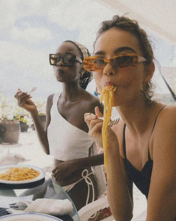 Two women in sunglasses eating pasta on a sunlit rooftop, mid-laugh, mid-bite — the long lunch as ritual.